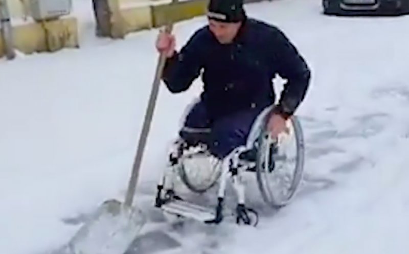 Bărbat în scaun cu rotile, cu picioarele amputate, surprins dezăpezind un trotuar